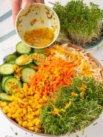 Oil-free garlicky dressing being poured over a bowl of alfalfa sprouts salad with corn, cucumber, carrot and shredded apple