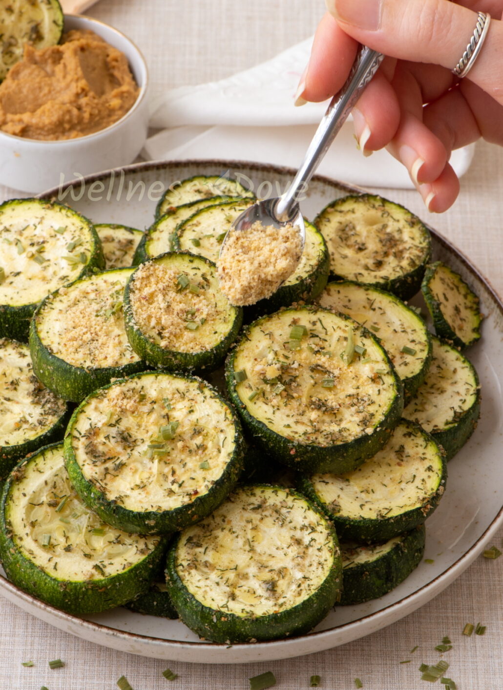 A plate full of roasted zucchini and a hand is sprinkling vegan parmesan over them