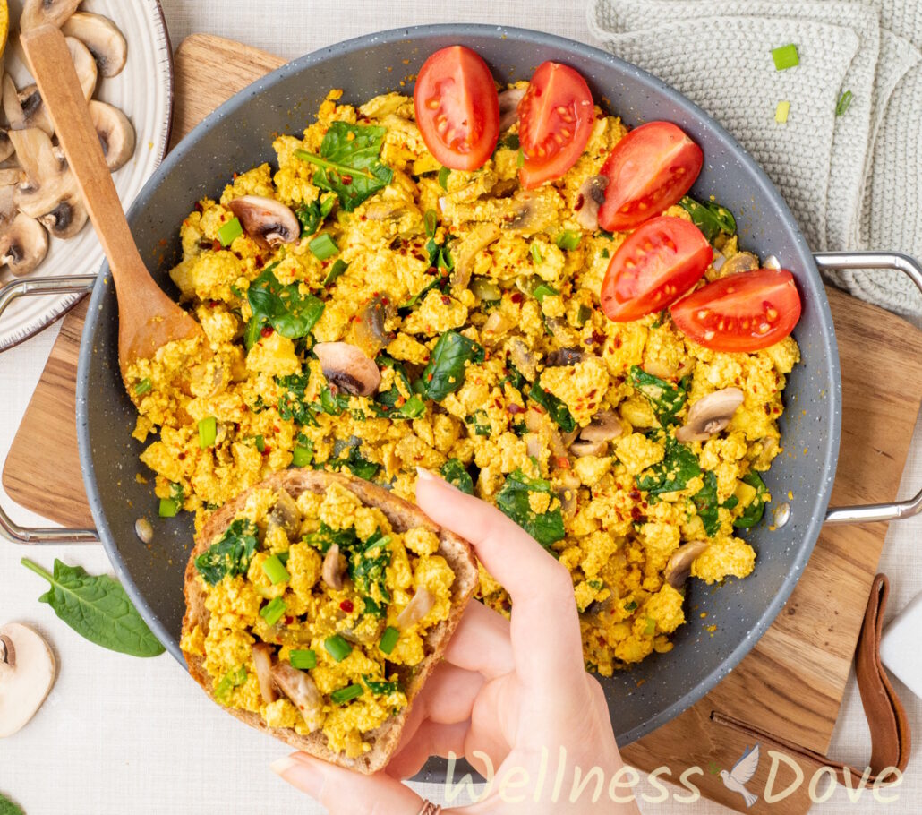 an overhead view of the Vegan Tofu & Spinach Egg Scramble in a pan while a hand is holding a sandwich over it
