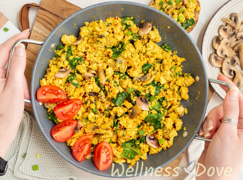 an overhead view of the Vegan Tofu & Spinach Egg Scramble in a pan while two hands are holding the handles of the pan