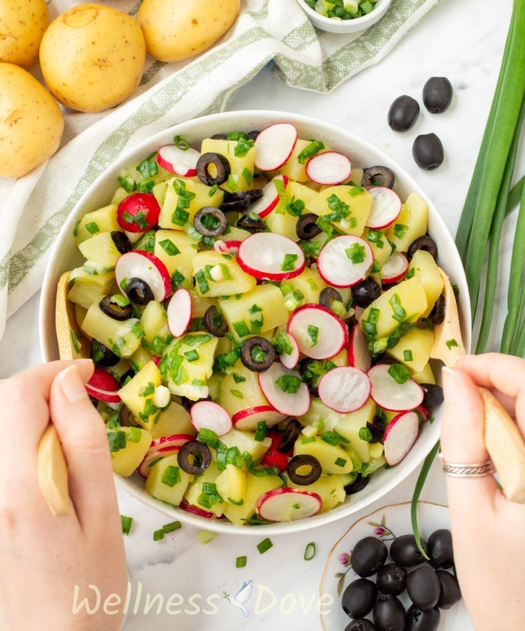 overhead view of the vegan no mayo potato salad , two hands are tossing the salad with wooden spoons