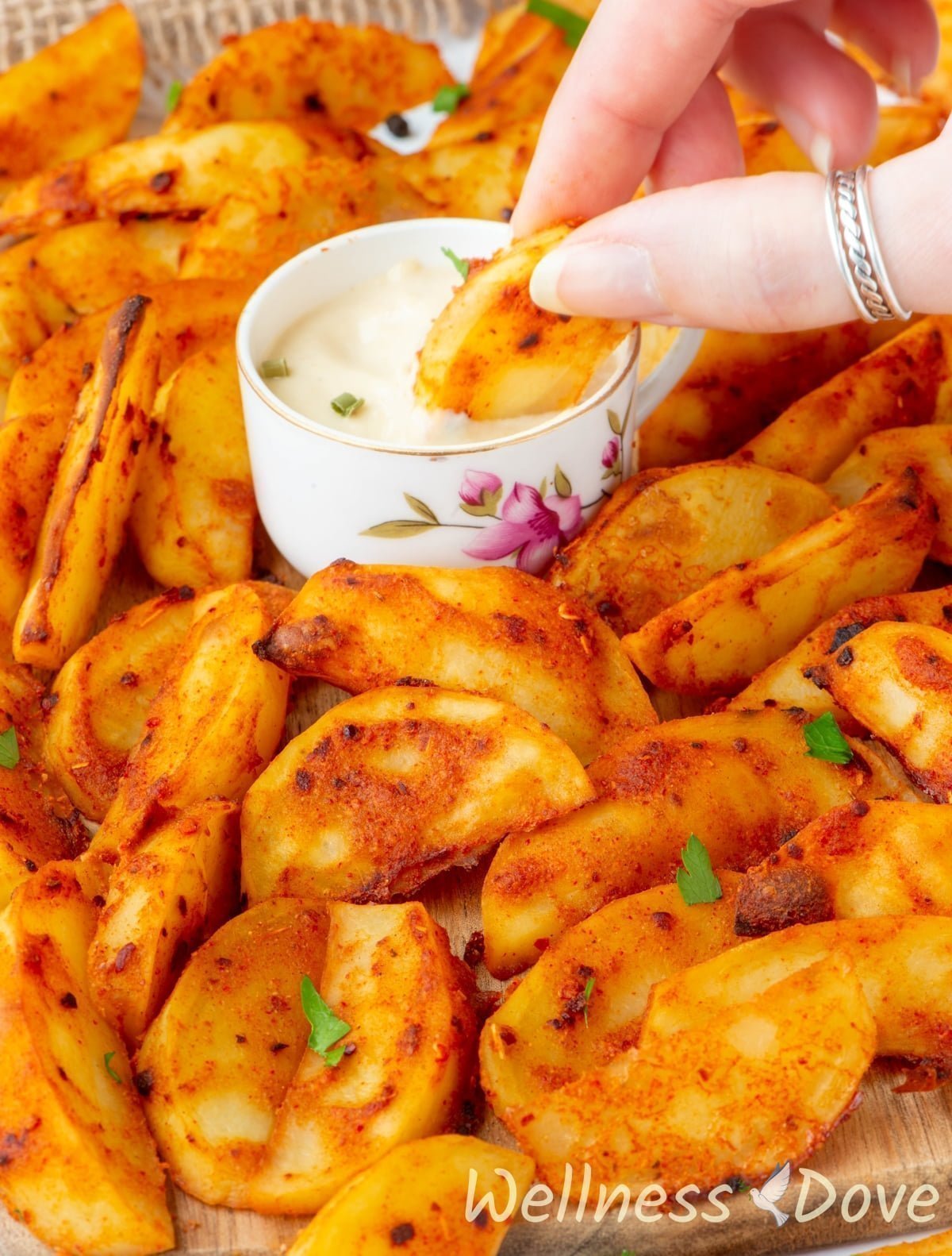 the Baked Mustard Potato Vegan Wedges on a chopping board and a hand is dipping one in cashew sauce